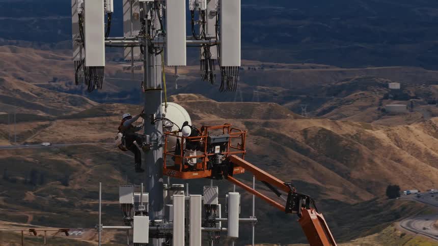 Cinematic aerial footage captures two technicians performing maintenance on a large telecommunications tower surrounded by rolling hills