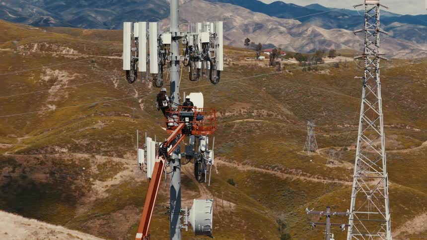 An aerial shot shows two technicians working on a tall cellular communications tower amidst a vast, hilly landscape