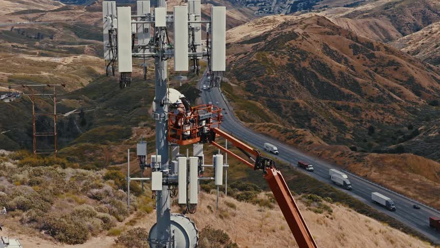 Aerial shot of two engineers on a crane working on a large cell phone tower overlooking a busy highway in a mountain pass