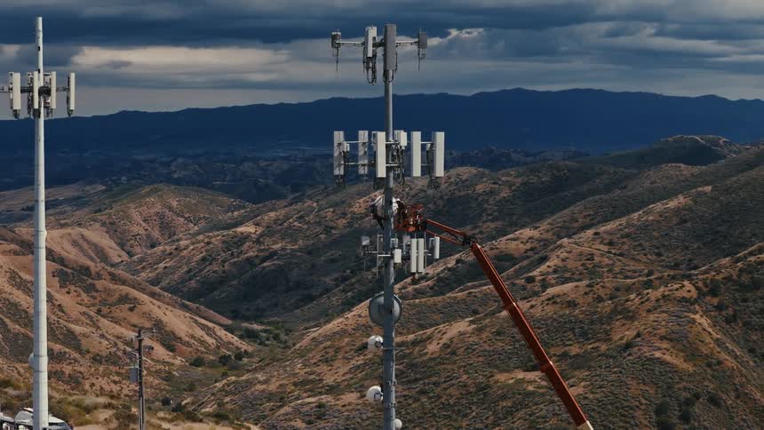An aerial view shows a technician in a crane basket working high on a cellular tower, set against a vast mountain landscape under an overcast sky