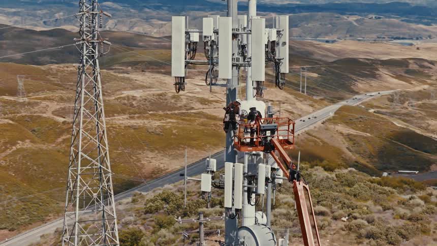 An aerial view shows two technicians in a crane performing maintenance high up on a cell tower above a rugged mountain landscape and busy highway