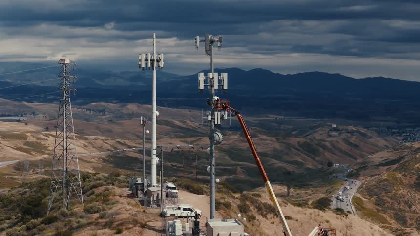 An aerial view shows a technician in a cherry picker performing maintenance on a telecommunications tower on a remote hilltop under a cloudy sky