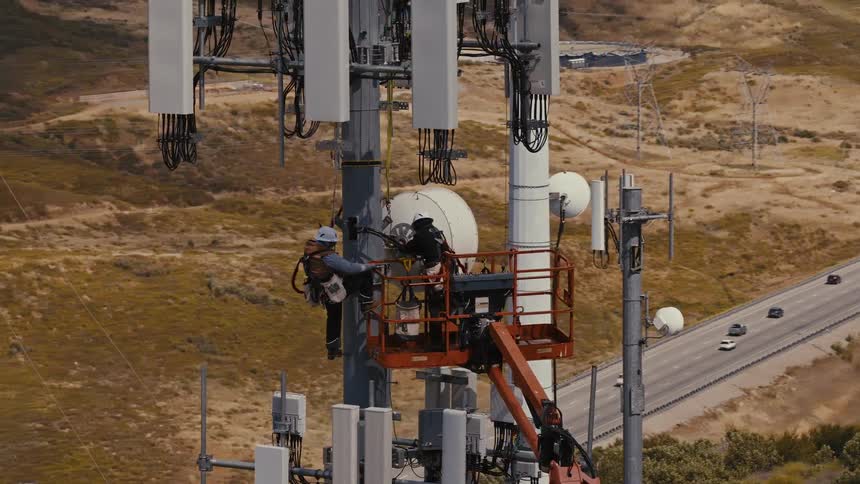 Aerial view of two skilled technicians in safety gear performing maintenance on a large telecommunications tower with a highway in the background