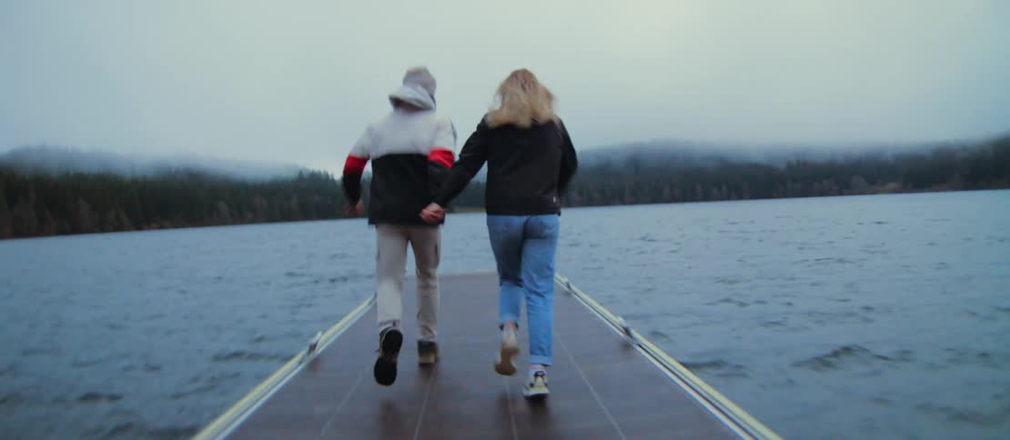 A happy couple runs down a dock holding hands on a foggy day, and the man picks up the woman and spins her around in slow motion
