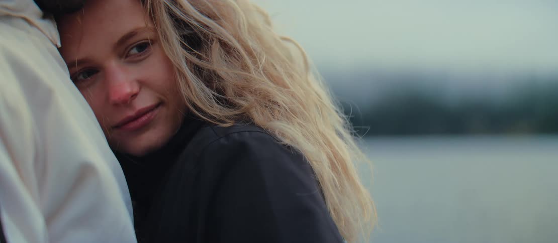 Close-up of a content young woman with curly blonde hair resting her head on her partner's shoulder by a lake