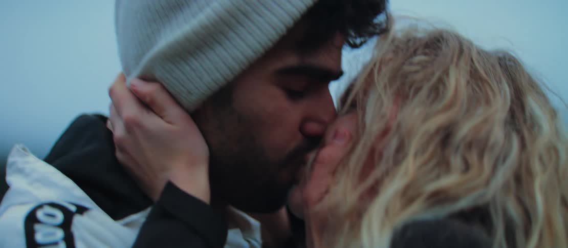 A romantic man proposes to his girlfriend by a lake on a cool day, placing the engagement ring on her finger as they share an emotional kiss and hug