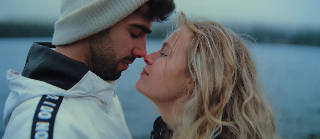 An intimate close-up of a young man and woman sharing a tender moment, nose to nose, against a serene watery backdrop