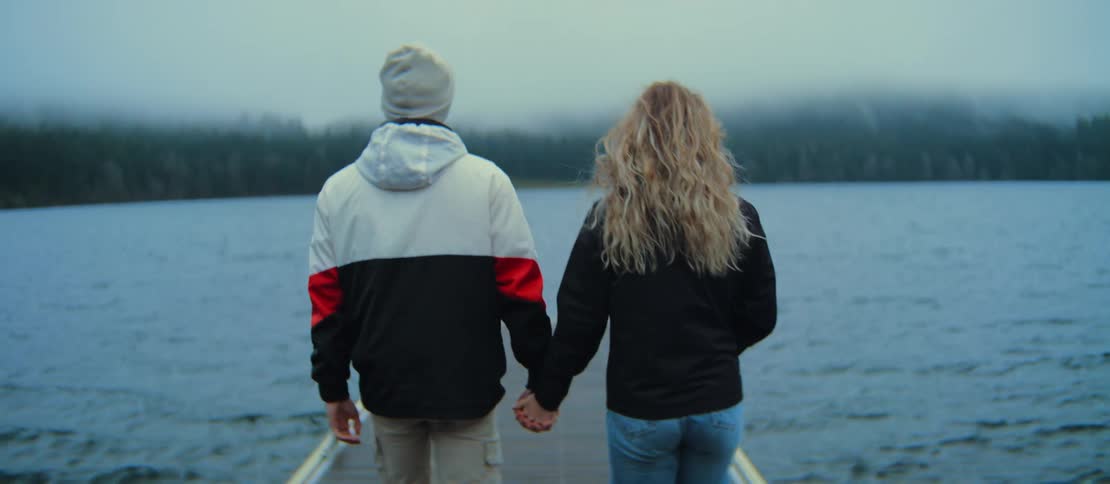 A young couple holds hands and walks together in slow motion down a wooden dock towards a vast, foggy lake on a cool day