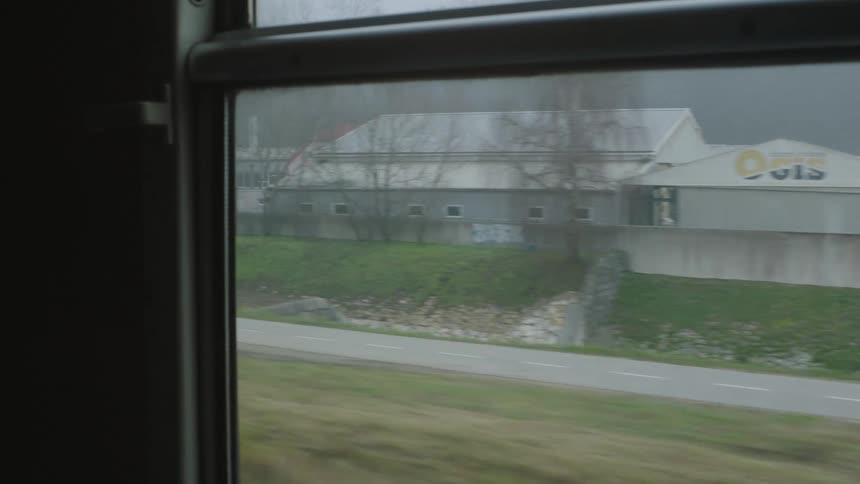 A steady tracking shot from a train window captures the passing scenery of an industrial town on a foggy and overcast day