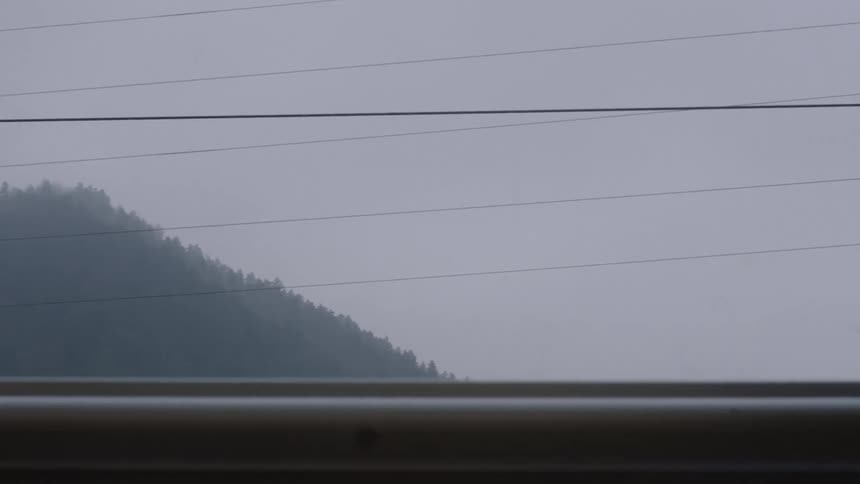 A point-of-view shot from a moving train window shows a foggy, tree-covered mountain on a gray, overcast day