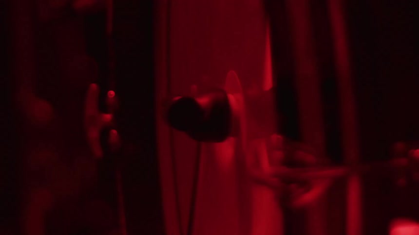 A moody close-up of earbuds hanging in a dark studio with dramatic red lighting as a musician plays an instrument in the background