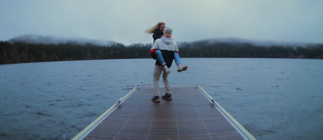 A man playfully spins his partner in his arms at the end of a dock on a foggy, tree-lined lake