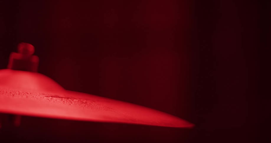 A drumstick strikes a cymbal in slow motion, sending a spray of water flying under a dramatic red light