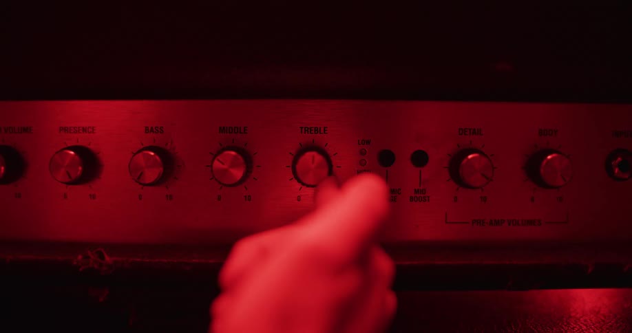 A hand adjusts the treble knob on a guitar amplifier under dramatic red lighting in a music studio