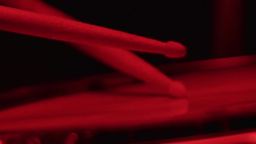A close-up of drumsticks playing a rhythm on a drum in slow motion, illuminated by a dramatic red light