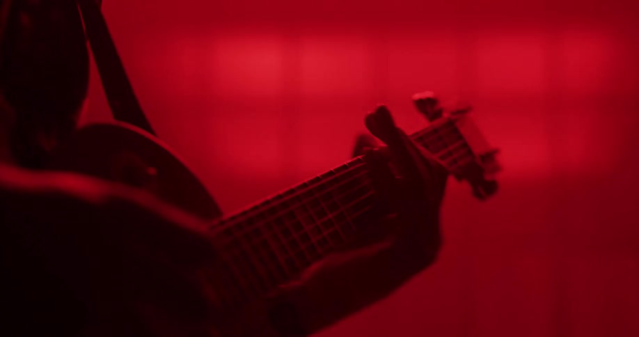 A silhouetted musician plays an electric guitar in a hazy room drenched in dramatic red light as the camera slowly pushes in