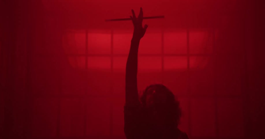 A drummer's hand twirls a drumstick in slow motion, revealed as a full silhouette at a drum set in a hazy, red-lit room