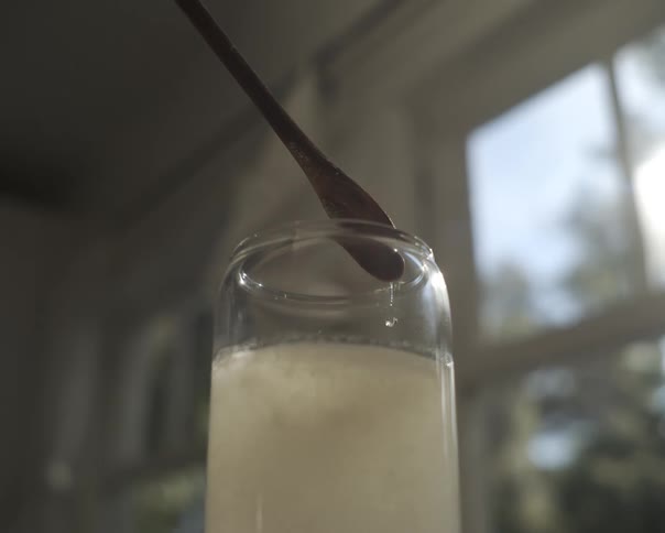 A close-up, slow-motion shot shows a hand lifting a small spatula out of a cloudy white liquid in a glass beaker, as droplets fall back into the mix