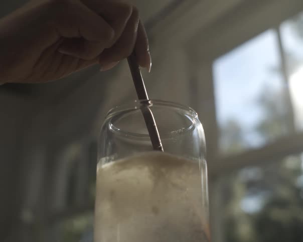 An artist's hand slowly mixes a substance into a glass jar of water with a paintbrush handle in slow motion