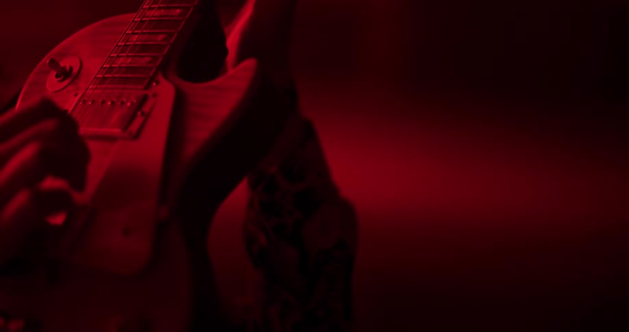 A tattooed musician skillfully plays a rock riff on an electric guitar in a dark room illuminated by intense red light