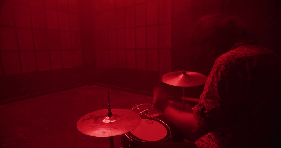A drummer energetically plays a drum kit in a dark, smoky room illuminated by intense red light