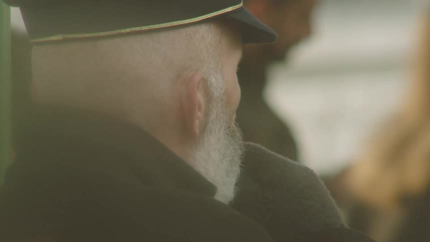 A contemplative, soft focus over-the-shoulder view of an elderly man with a white beard in a hazy public space