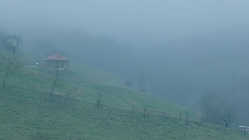 Tracking shot through a foggy window reveals a mysterious rural landscape with a lone gazebo perched on a green hill
