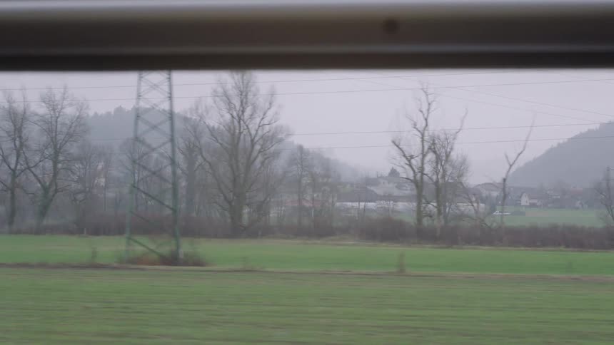 Passing by a green countryside with a foggy mountain backdrop as seen from a moving train on a gloomy day