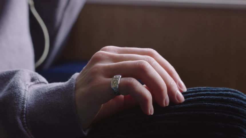 A close-up of a person's hand wearing a silver ring resting on their knee as their fingers gently twitch