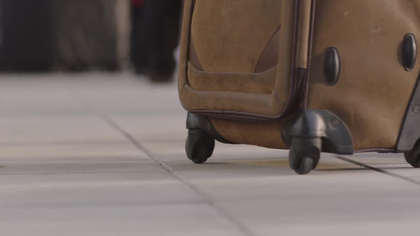 A slow-motion, low-angle rack focus shot shows a traveler pulling a rolling suitcase across a terminal floor