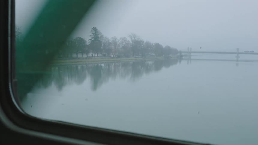 Passing a calm river and a foggy landscape with a distant bridge, as viewed from a train window