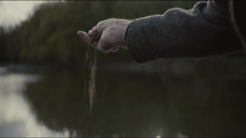 A person's hand slowly opens to release and scatter sand or ashes over a tranquil lake in slow motion