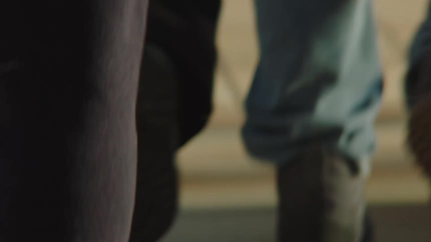 A close-up, low-angle shot follows the feet and legs of a crowd of people walking through a public space, some with luggage