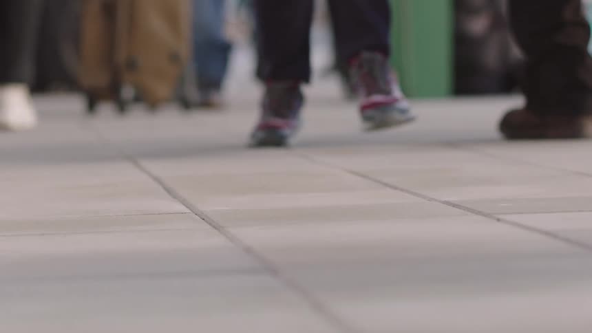 A low-angle super slow-motion shot of a crowd's feet walking away from the camera on a tiled floor