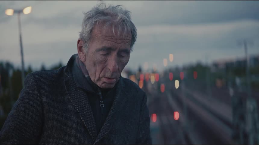 Slow motion close-up of a somber elderly man smoking a cigarette on an overpass at dusk with city lights in the background