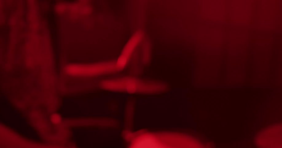 An energetic drummer performs a solo in a dark, moody room bathed in intense red light, captured with blurry, artistic shots