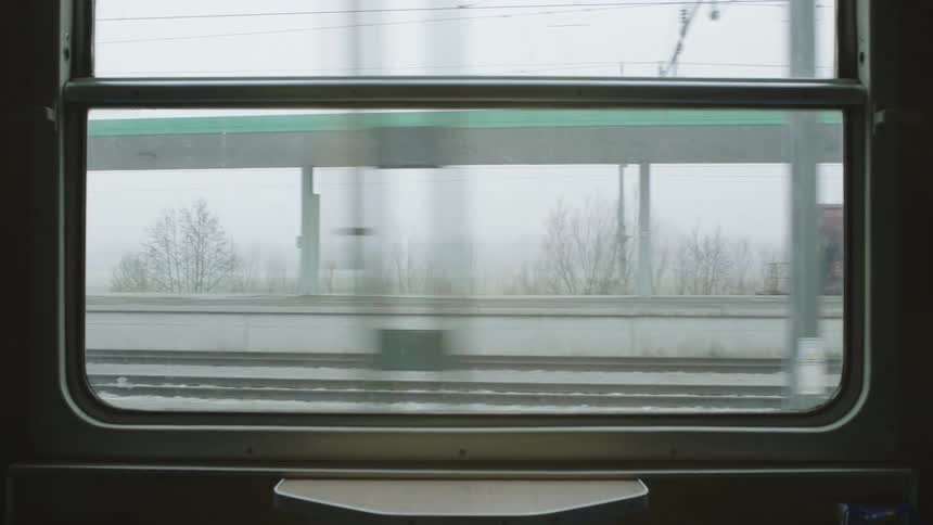 An interior view from a moving train shows a freight train passing by the station on a gray, misty day