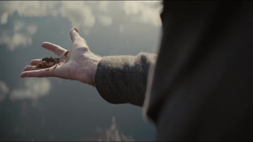 A man lets soil slip through his fingers over a tranquil lake reflecting the cloudy sky in slow motion