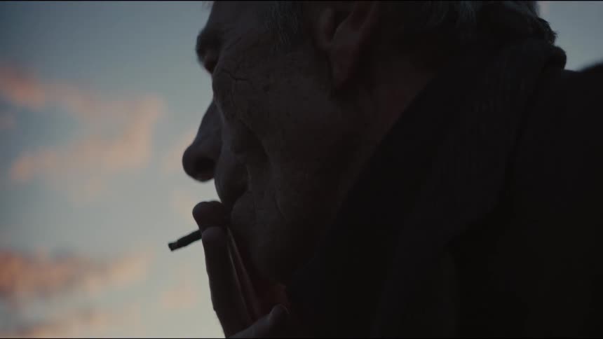 An elderly man in silhouette thoughtfully smokes a cigarette against the dusky evening sky in slow motion