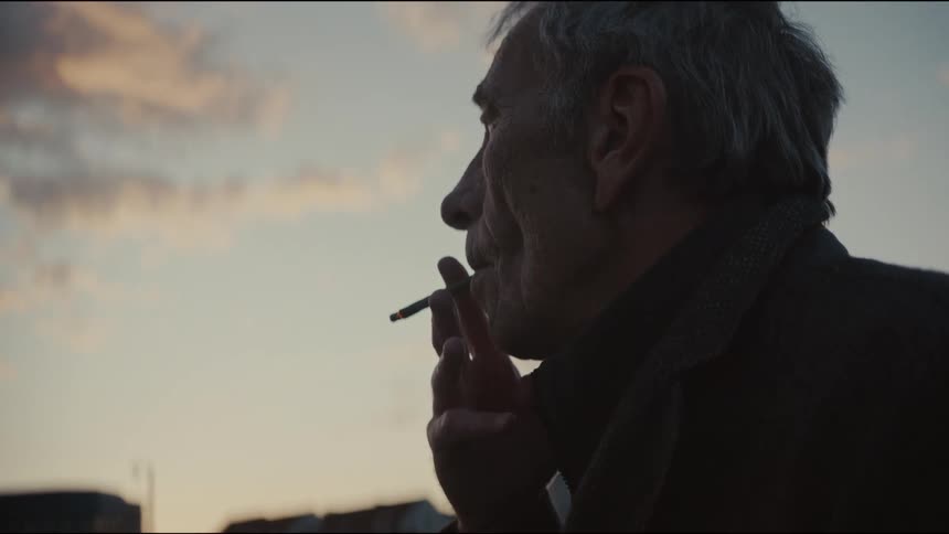 A contemplative older man smokes a cigarette in slow motion while watching the sunset over the city