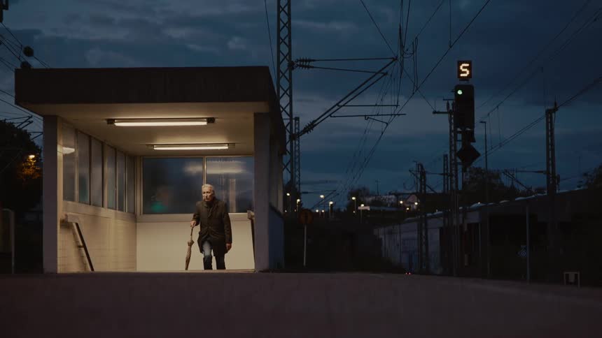 An elderly man with an umbrella slowly walks up the stairs to a deserted train platform at night