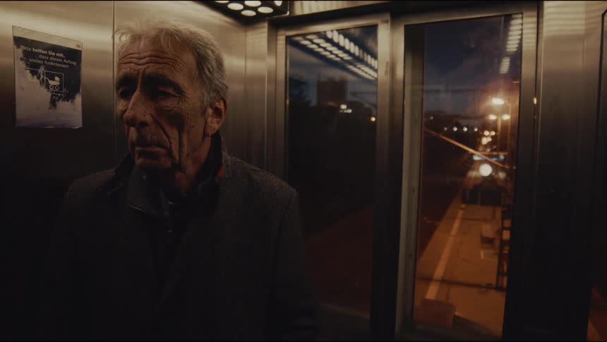 A weary older man stands pensively in a modern elevator as the doors close, revealing the lights of the train station and city at night