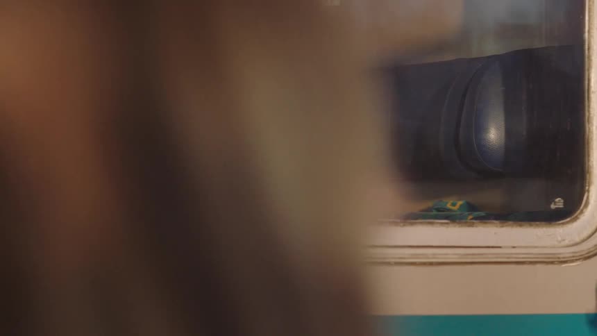 A slow pan across a train car window reveals various passengers inside during their journey