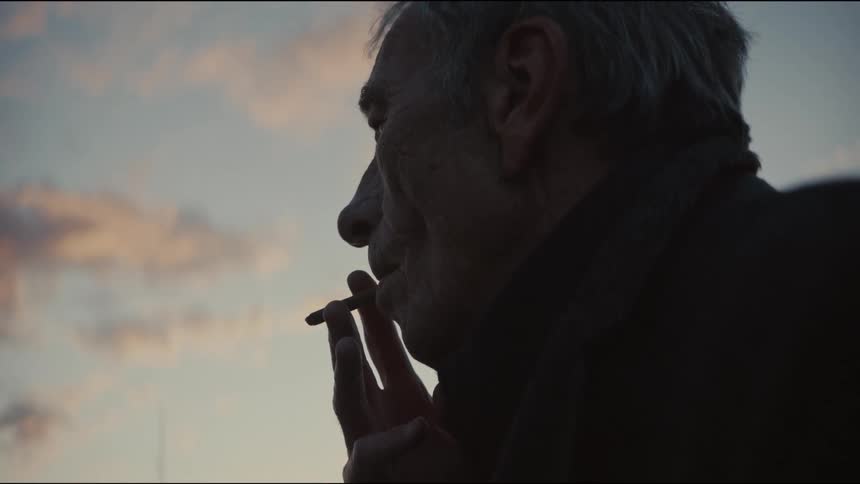 A silhouetted elderly man smokes a cigarette in slow motion against the twilight sky