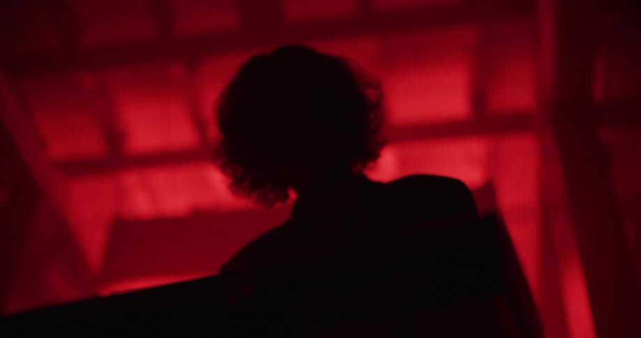 A silhouetted musician with curly hair grooves in slow motion in a dark room bathed in red light