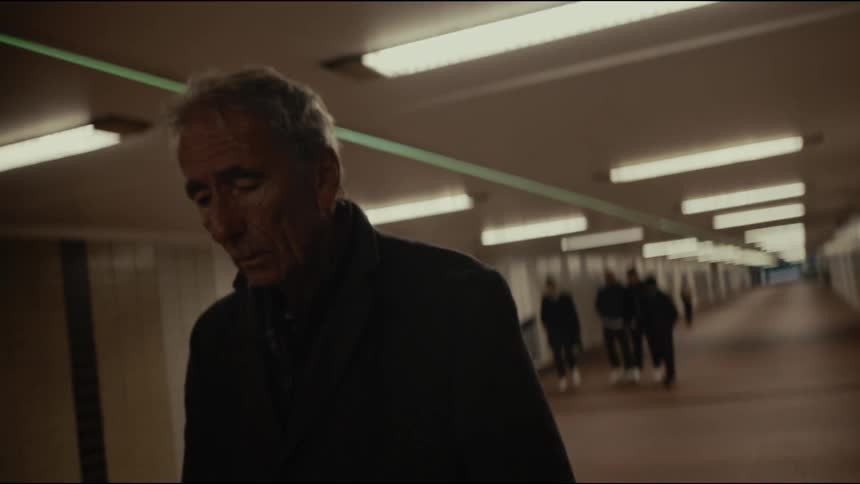 Slow-motion close-up of a somber elderly man walking through a dark pedestrian tunnel as a group of youths approaches