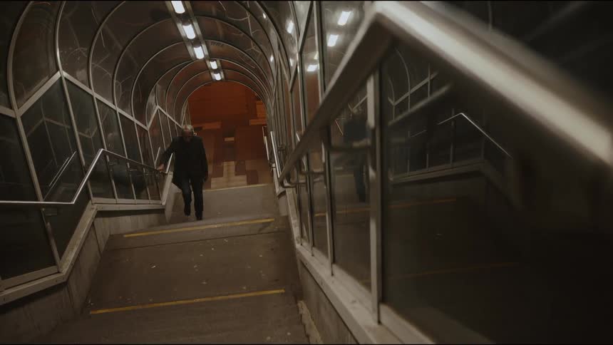 A lone man with long grey hair slowly climbs the stairs of a covered pedestrian tunnel at night in an urban setting