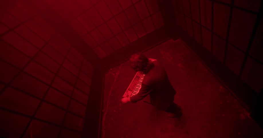 From above, a musician emotionally plays a synthesizer in a claustrophobic red room before ending his performance in frustration