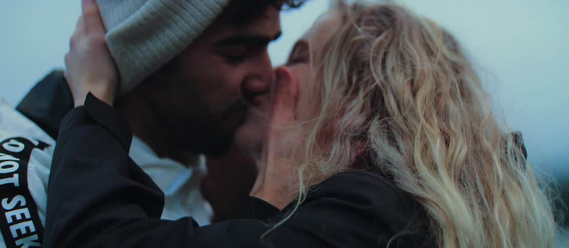 A young man in a white beanie proposes to his girlfriend outdoors, placing an engagement ring on her finger as they share a loving kiss and hug