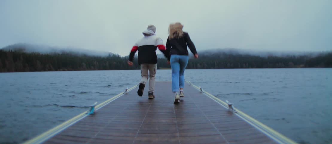 A playful young couple runs hand-in-hand in slow motion down a floating dock on a foggy lake, culminating in a joyful, spinning embrace